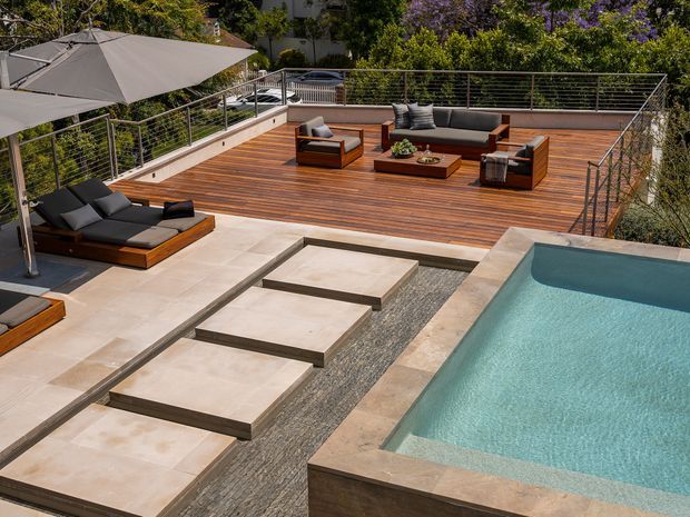 A contemporary outdoor entertaining area with a timber deck, low lounge seating, large umbrellas, a rectangular swimming pool, and a paved stepping-stone path set in a strip of dark gravel. Horizontal cable railings frame the elevated terrace, with landscaped greenery beyond.