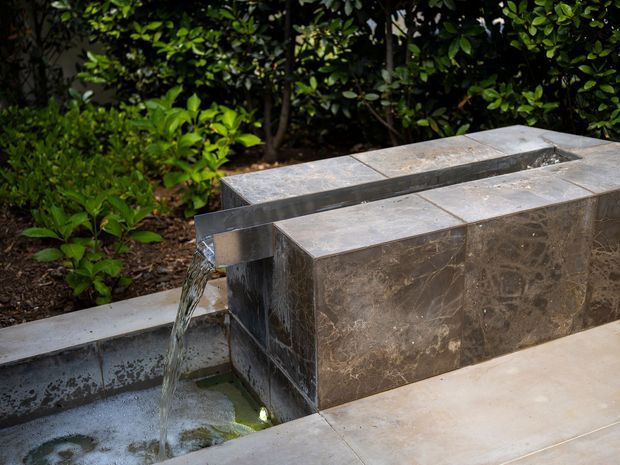 Close-up of a contemporary rectangular water feature set into a landscaped garden. Water flows from a narrow slot between dark stone-clad blocks into a shallow tiled basin, with dense green planting behind it and pale paving in the foreground.