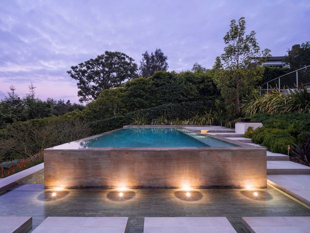 Twilight view of a landscaped residential pool area with a raised rectangular infinity-edge pool or reflecting pool. The pool is enclosed by a substantial warm-toned stone or concrete feature wall with integrated uplighting, surrounded by paved terraces, planted garden beds, clipped hedges, and steps leading toward a partially visible contemporary home.