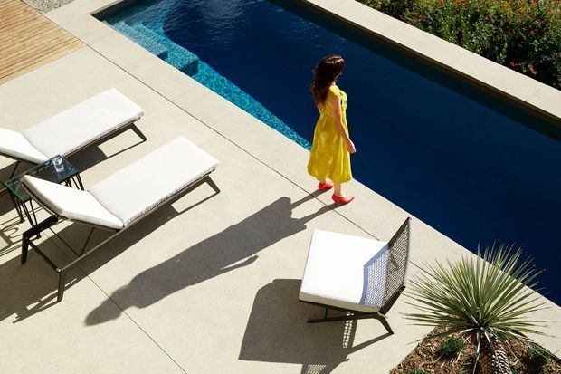 A sunlit modern pool terrace viewed from above, with a long rectangular dark-blue swimming pool edged in pale concrete, white cushioned lounge chairs, a timber deck, and drought-tolerant planting. A person in a bright yellow dress walks along the pool edge.