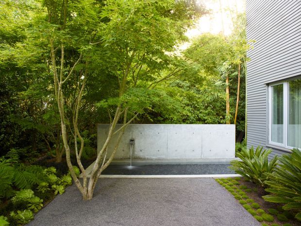 A landscaped side courtyard beside a modern building, featuring a smooth grey rendered or concrete privacy wall with a narrow water spout, dark linear water feature, gravel or exposed-aggregate paving, lush established greenery, and a contemporary grey horizontal-clad facade with a large window.