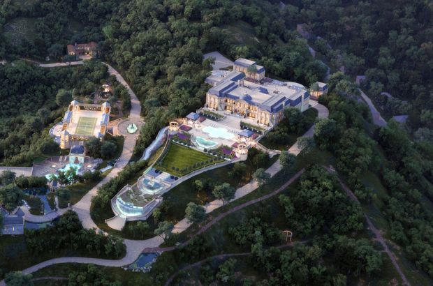 Aerial view of an expansive luxury estate set within dense, heavily landscaped woodland. The complex includes a large formal main building, multiple outdoor terraces, swimming pools, ornamental water features, manicured lawns and gardens, winding private paths, and a separate sports-court pavilion. The architecture appears grand and classically detailed, with pale stone façades, dark pitched roofs, and illuminated interior spaces.