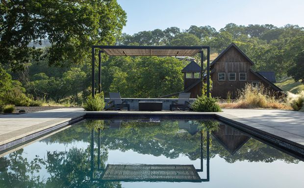 A tranquil outdoor pool area set within a lush wooded landscape. A dark rectangular swimming pool reflects a slim pergola with a slatted roof, positioned beside a paved terrace with lounge chairs and ornamental grasses. Behind it is a rustic dark timber-clad building with steep gabled roofs and small square windows, partially integrated into the surrounding greenery.