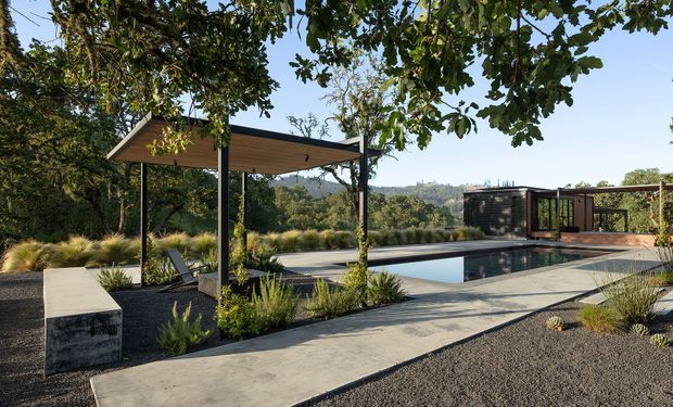 A landscaped outdoor pool area with a long rectangular swimming pool, concrete paths, dark gravel garden beds, native grasses and mature trees. A simple open-sided pavilion with a timber-lined flat roof provides shade beside the pool, while a dark-clad pool or garden structure and timber deck sit beyond it. The setting appears elevated or sloped, with wooded hills visible in the distance.