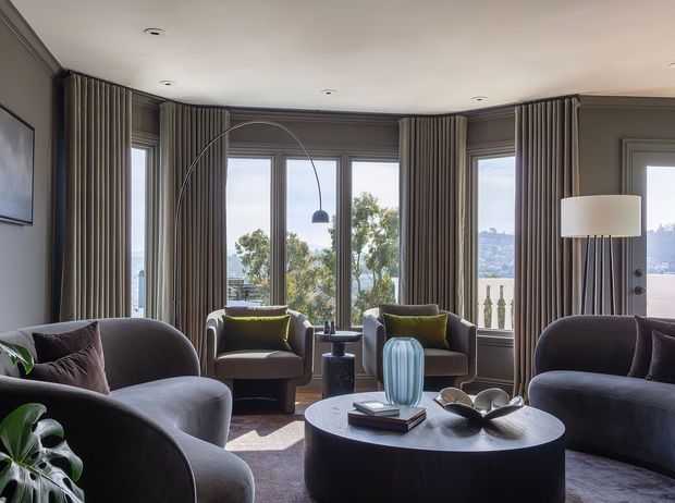 Refined living room with curved upholstered lounge chairs and sofas arranged around a large round coffee table. Floor-to-ceiling bay windows are dressed with full-height striped curtains, while a sculptural arched floor lamp, table lamp, and decorative accessories add visual interest. The room has dark grey walls and furnishings, soft natural light, and views toward trees and a hillside beyond the windows.