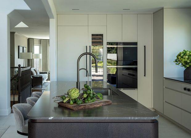 A refined contemporary kitchen with a large dark stone island, integrated pale cabinetry, black hardware, and a sculptural dark tap. Upholstered bar stools line the island, while an open-plan living area is visible beyond. The kitchen features full-height joinery, integrated appliances, a reflective splashback or glass panel, and soft natural light.