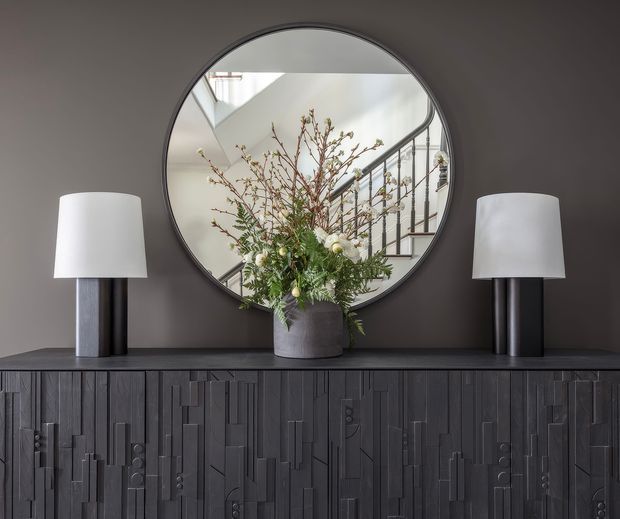 A refined interior vignette featuring a dark textured sideboard with sculptural geometric detailing, two matching table lamps, and a large round mirror. The mirror reflects a bright stairway and railing, while a large floral arrangement in a grey vessel provides an organic focal point. The palette is predominantly charcoal, grey, white, and muted green, creating a sophisticated, moody atmosphere.