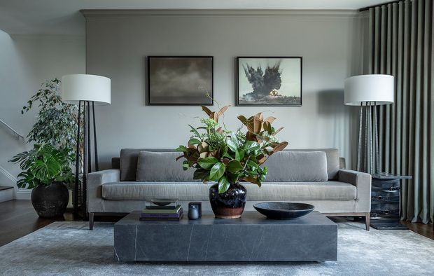 A refined living room with a long grey upholstered sofa, dark marble or stone rectangular coffee table, tall floor lamps, layered grey curtains, framed artwork, and large indoor plants. The room has a muted neutral palette, tailored built-in wall panelling or moulding, and a calm contemporary-classic feel.