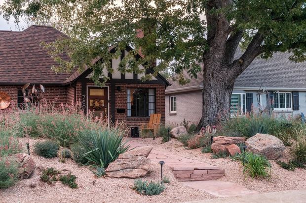 Front exterior view of a low-rise brick house with a dark shingle hipped roof, small covered entry, and adjacent residential structures. The landscaped front garden features xeriscaping with gravel, large natural boulders, native-style grasses, spiky architectural plants, and a stone-paver path leading to the entrance. A mature tree dominates the foreground.