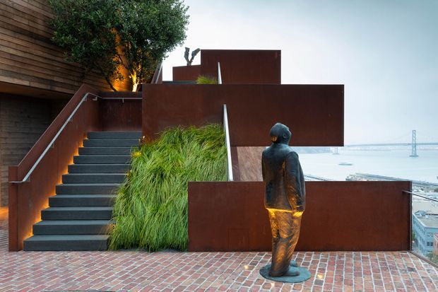 A contemporary rooftop or terrace setting with dark rusted steel privacy screens, a wide external staircase, dense ornamental grasses and a sculptural figure. Red-brown brick paving covers the terrace, while a distant waterfront or harbour is visible beyond the building edge.