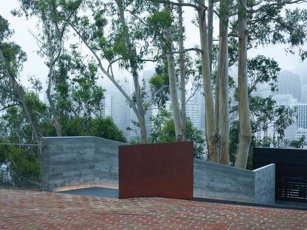 A landscaped rooftop or terrace with red brick paving, low raw concrete walls, and a rust-coloured metal privacy screen. Mature eucalyptus trees frame views toward a dense urban skyline in the background. A dark slatted structure or gate is visible at right, creating a contemporary outdoor architectural setting.