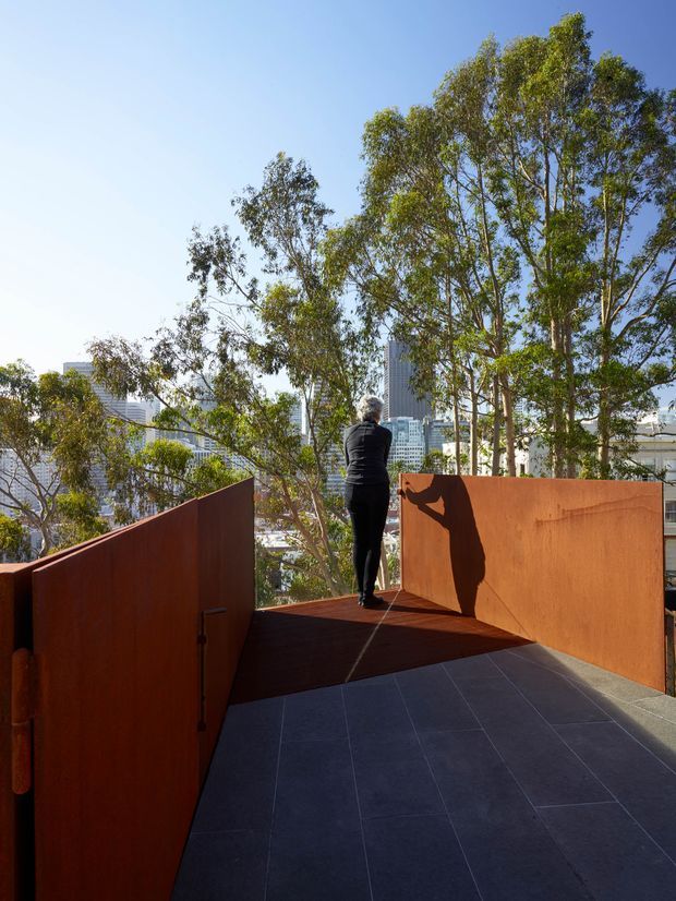 A sunlit elevated outdoor terrace or walkway with dark large-format tiled paving and tall rust-coloured steel privacy screens. A person stands at the end of the terrace, overlooking mature trees and a dense urban skyline. The image emphasizes the sculptural ramp-like circulation space, warm weathered metal surfaces, strong shadows, and city views.