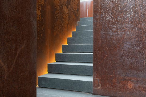 A dramatic interior stairway with broad dark grey stone steps ascending between tall rusted steel or weathered metal wall panels. Warm concealed lighting glows along the sides of the stairs, creating a strong contrast with the cool stair surfaces. The image is tightly framed and emphasizes the materiality, texture, and sculptural character of the stair enclosure.