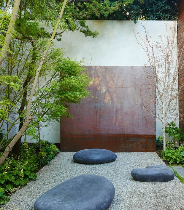 A secluded landscaped courtyard garden with a pale rendered boundary wall and a large rusted weathering-steel feature panel. Fine gravel covers the ground, punctuated by smooth dark stone-like sculptural stepping forms. Layered green planting and slender ornamental trees frame the space, with part of a timber-clad building visible at the right edge. The composition is minimalist and serene, combining natural planting with sculptural materials.