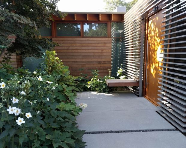 A landscaped courtyard or entry garden with a contemporary timber-clad building, warm wood-framed glazing, and a striking horizontal metal louvre screen enclosing a timber door. A floating timber bench projects from the screened wall, while lush flowering planting borders a smooth concrete-paved path. The scene has a carefully composed, modern indoor-outdoor character with dappled sunlight.
