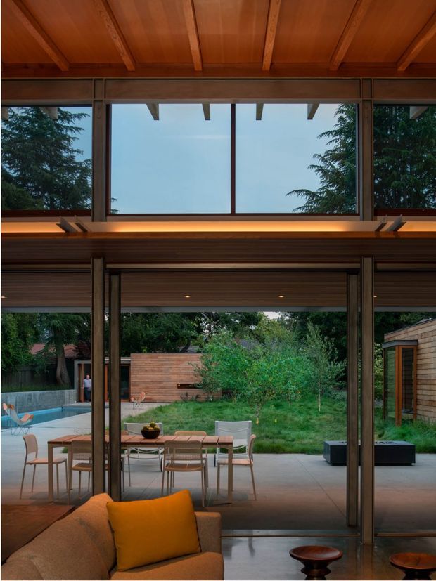 Interior view of a warm timber-framed living room opening onto a landscaped courtyard and pool terrace. Full-height glazed sliding doors, exposed timber ceiling beams, clerestory glazing, and a broad timber-lined transition detail connect the living area with the outdoor dining and garden spaces. Neutral seating and dining furniture create a relaxed, contemporary atmosphere.