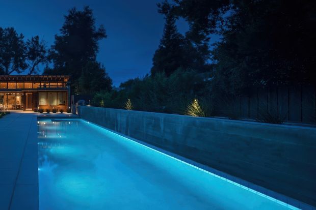 Twilight exterior view of a long illuminated swimming pool beside a paved terrace and tall retaining wall, with landscaped planting and a warmly lit modern pavilion or house at the far end. Dense mature trees surround the property, creating a secluded, atmospheric setting.