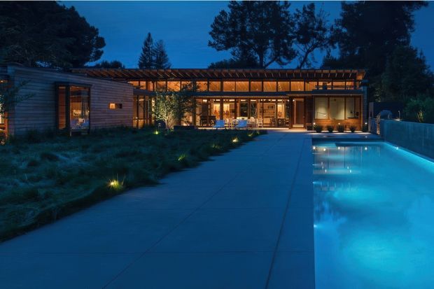 Twilight exterior view of a low, contemporary residential pavilion with extensive warm-lit glazing, horizontal timber-clad walls, a pergola-like roof structure, landscaped planting, broad concrete paving, and a long illuminated swimming pool in the foreground. Mature trees frame the property.