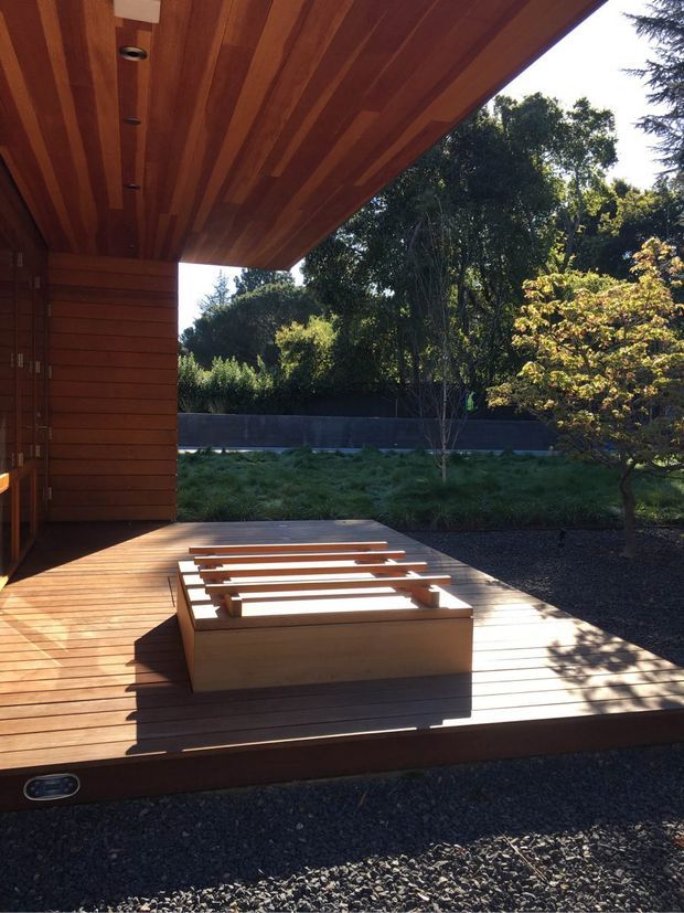 A timber-clad covered outdoor terrace or deck beside a landscaped garden. The space features warm timber decking, a timber-lined soffit and wall, and a substantial built-in timber bench or platform with slatted detailing. Strong sunlight creates pronounced shadows across the deck, while mature planting and a dark boundary wall frame the view.