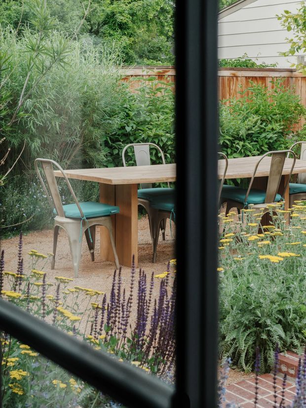 View through dark-framed glazing onto a landscaped outdoor dining area. A rectangular timber table is surrounded by green-cushioned metal chairs, with dense planting, flowering perennials, a timber fence, and a neighbouring light-clad wall beyond. Brick paving is visible in the foreground.