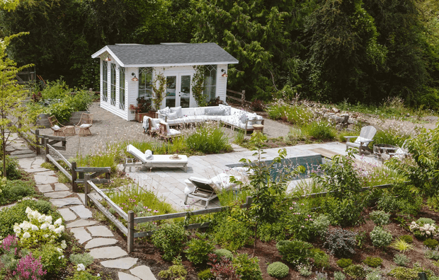 A landscaped garden retreat featuring a small white glazed pavilion or pool house, a paved outdoor entertaining area with light-colored lounge seating, a partially visible swimming pool, and informal stone pathways surrounded by dense planting and mature trees.