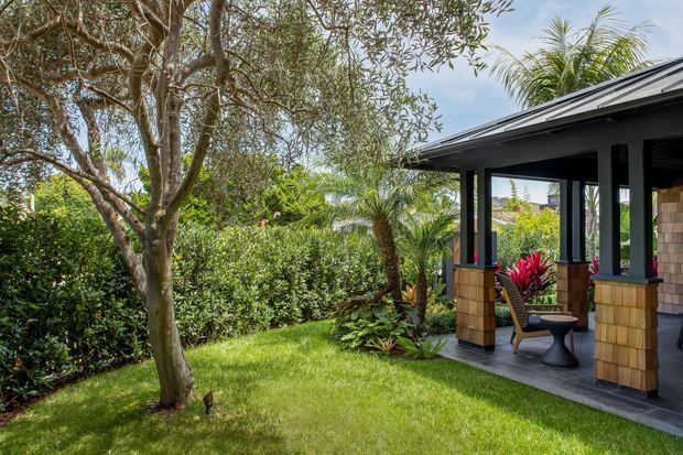 A lush landscaped garden beside a covered outdoor pavilion or veranda. The dark-framed structure has a pitched metal roof, substantial square posts with warm shingle-clad bases, dark tiled flooring, and outdoor seating. Dense tropical and subtropical planting surrounds the lawn, creating a private garden setting.