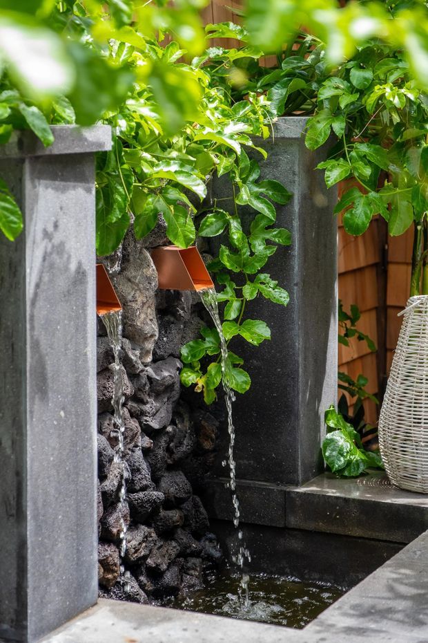 A lush outdoor garden water feature with dark stone or concrete pillars framing a cascading rock fountain. Water flows from small copper-toned spouts into a shallow black-lined basin, surrounded by dense glossy green foliage and timber screening. A woven decorative lantern or basket sits on the ledge to the right.