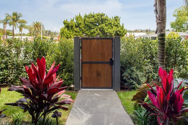 A landscaped garden walkway leads to a contemporary timber pedestrian gate set within dark masonry or metal side panels. Lush hedges and vivid red tropical foliage frame the entrance, with palm trees and a bright suburban setting beyond.