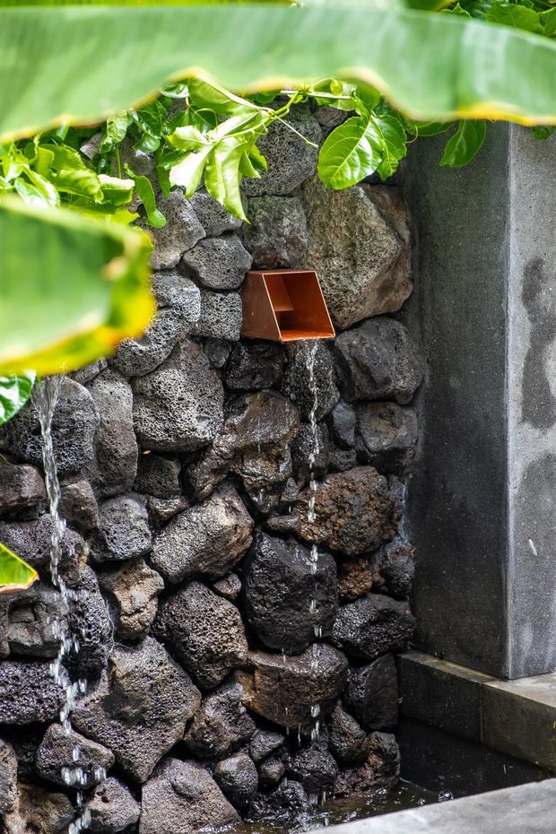 Close-up of a landscaped water feature with water flowing over a wall of dark porous volcanic rocks. A small rust-coloured metal spout projects from the stonework, with lush green leaves framing the top and a dark concrete or stone edge and basin visible at the right and bottom.