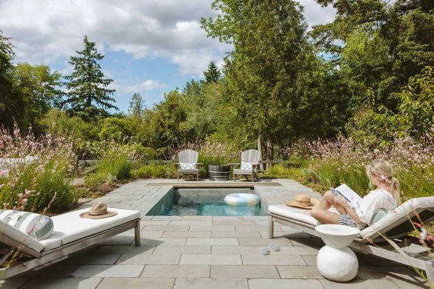 A landscaped outdoor patio with a rectangular in-ground swimming pool, surrounded by pale stone paving, lounge chairs, and lush flowering garden planting. The scene is bright and relaxed, with a person reclining on a sun lounger and additional seating at the far end of the pool.