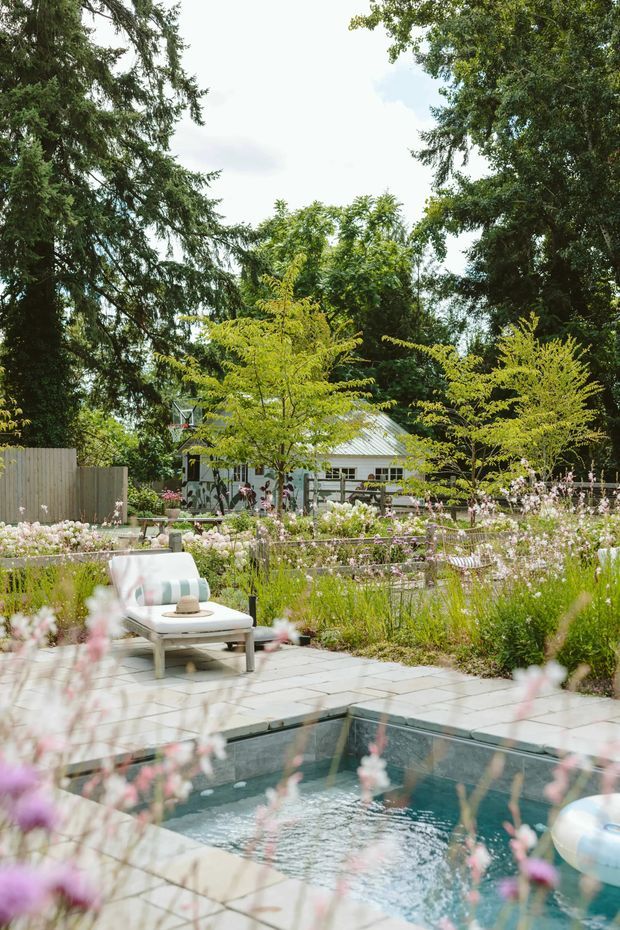 A landscaped residential garden with a small in-ground pool in the foreground, pale stone paving, a white outdoor lounge chair, abundant flowering plants, mature trees, and a light-colored garden pavilion or house in the background. The scene has a relaxed, natural, resort-like atmosphere.
