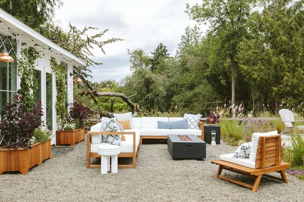 A landscaped outdoor lounge and patio beside a light-coloured glazed garden room or house extension. The gravel surface is furnished with a pale outdoor sofa, timber-framed armchairs, a low dark coffee table, and a timber sun lounger, surrounded by mature planting and climbing greenery.