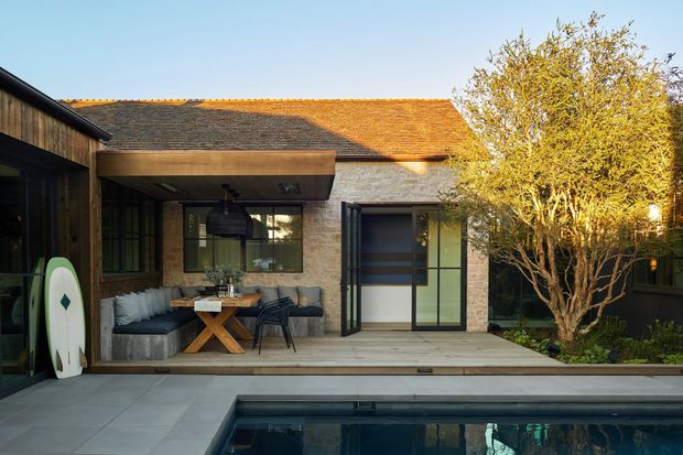 A contemporary courtyard-style residential exterior with a covered outdoor living area opening onto a paved terrace and swimming pool. The building combines dark timber cladding, pale brick walls, large glazed doors and windows, and a pitched tiled roof. Outdoor lounge furniture, a surfboard, planted landscaping, and a sculptural tree create a relaxed, warm setting.