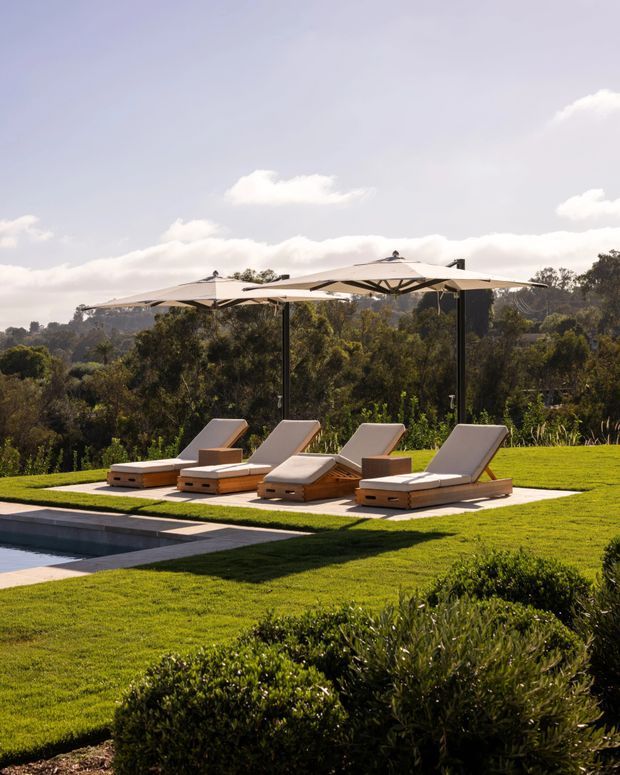 A landscaped outdoor poolside area with four cushioned sun loungers arranged on a pale paved terrace beneath large cantilevered umbrellas. The setting includes manicured lawn, clipped shrubs, a swimming pool edge, and a wooded hillside beyond, creating a refined resort-like outdoor space.