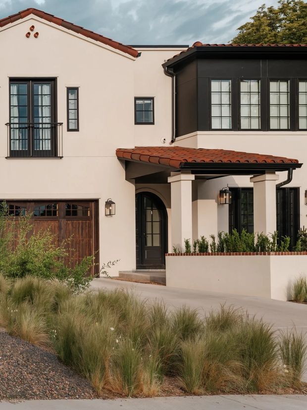 Exterior view of a refined two-storey residence with cream stucco walls, dark-framed multi-pane windows, terracotta tiled gable and porch roofs, a covered entry supported by square columns, and a landscaped frontage with ornamental grasses. The facade combines traditional Mediterranean-inspired detailing with a darker contemporary glazed projecting section.
