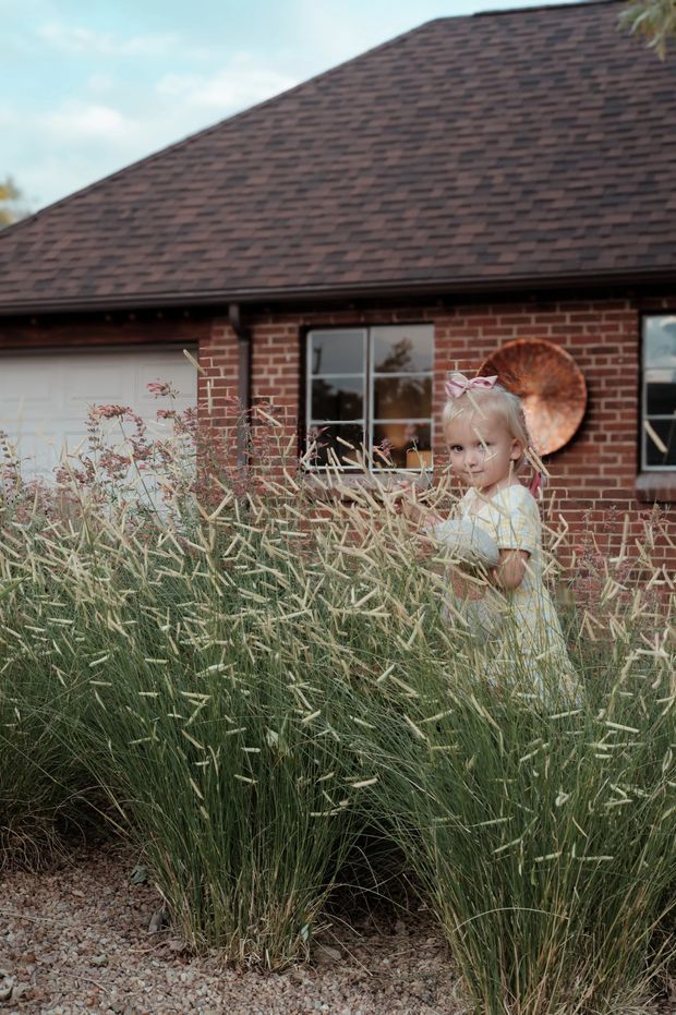 A residential brick house viewed from a landscaped front garden, with a dark shingled pitched roof, white garage door, black-framed windows, and tall ornamental grasses in the foreground. A young child is partially visible among the planting. The image has a warm, rustic suburban character.