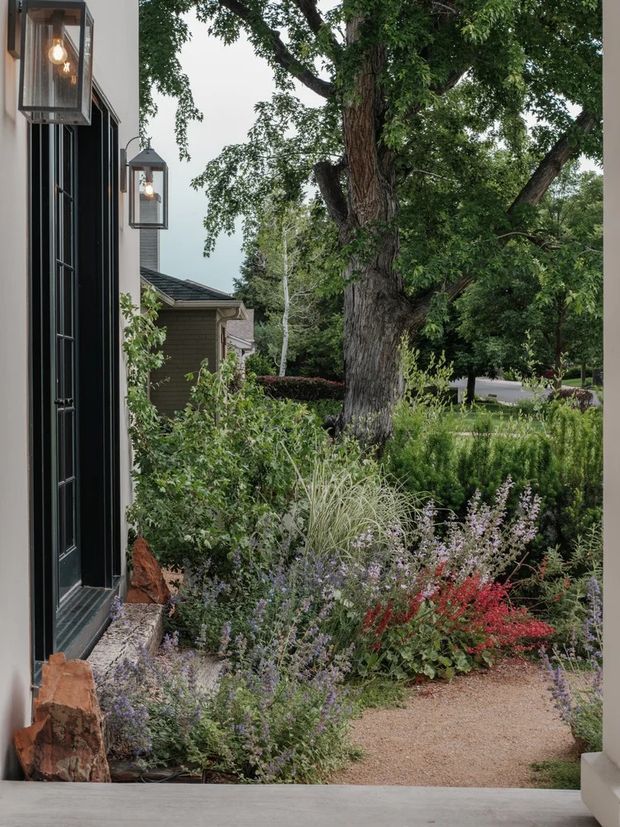 A landscaped residential entry viewed from a covered threshold, with a dark paneled exterior wall and glazed door at left, suspended lantern sconces, a gravel garden path, layered ornamental planting, and a mature tree dominating the front garden. A neighbouring residential structure and street are visible beyond the garden.