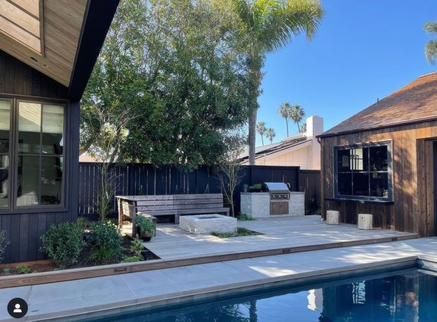 A contemporary outdoor pool courtyard beside dark timber-clad buildings. The space features a pale timber deck, rectangular swimming pool, built-in bench seating, a compact outdoor kitchen with barbecue, dark fencing, and lush tropical planting under a clear blue sky.