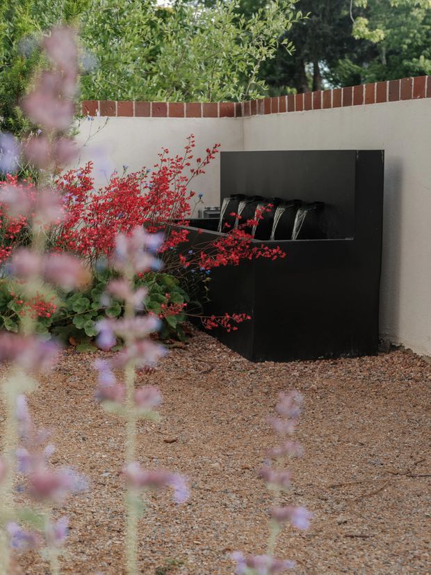 A landscaped outdoor courtyard corner featuring a black rectangular built-in water feature or fountain set against a light rendered wall topped with red brick coping. Gravel ground, mature greenery, and flowering plants surround the minimalist feature, creating a calm garden setting.