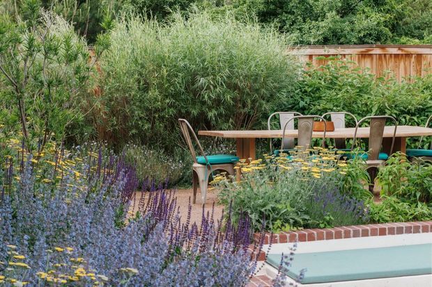 Lush landscaped outdoor garden beside a swimming pool, with abundant flowering and ornamental planting, a timber dining table and metal chairs, and a timber privacy fence in the background. A low brick-edged raised garden bed borders the pool, creating a colourful, natural outdoor entertaining setting.