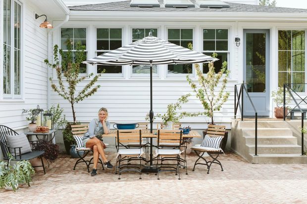 A bright residential courtyard patio beside a white weatherboard home, furnished with a small outdoor dining table, folding chairs, and a striped umbrella. The patio has pale herringbone paving, raised concrete steps, black metal railings, large multi-pane windows and French doors, with potted plants and climbing greenery adding a relaxed outdoor feel.