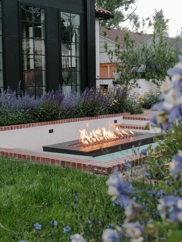 A landscaped residential courtyard with a built-in linear gas fire feature set into a sunken tiled terrace. Red brick edging, pale rendered walls, dark stone or concrete fire surround, and abundant lavender-like planting frame the outdoor seating area. Behind it is a dark, finely gridded glazed facade alongside a traditional pitched-roof building, creating a refined blend of contemporary and traditional elements.
