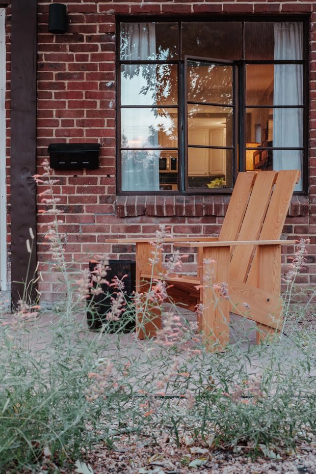 Exterior view of a brick building facade with a dark timber-framed multi-pane window, partially open casement, and warm interior visible through the glazing. A light timber Adirondack-style chair sits in a planted gravel garden beside the window, with a black wall-mounted mailbox and exterior light.