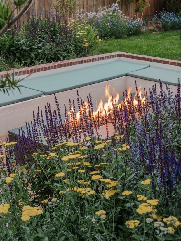 A landscaped residential garden featuring a sunken rectangular fire pit with a warm flame, pale rendered walls, and integrated pale green bench seating. A red brick coping border surrounds the pit, which is embedded among dense flowering plantings with purple and yellow blooms. Timber fencing and lawn form the background.