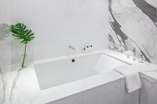 A bright, luxurious bathroom featuring a deep built-in white bathtub surrounded by white marble surfaces with dramatic grey veining. Chrome tapware, a folded white towel, toiletries, and a green tropical plant complete the clean spa-like setting.