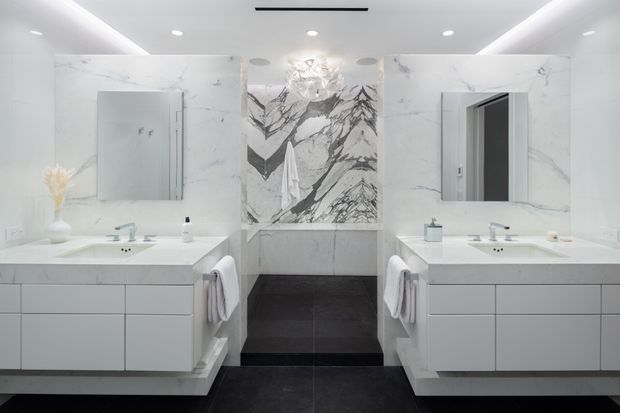 A bright, symmetrical contemporary bathroom with a double vanity, large frameless mirrors, white marble-look wall surfaces and benchtops, and dark charcoal floor tiles. A freestanding bathtub sits centrally against a dramatic veined marble feature wall, with recessed ceiling lighting and minimalist white cabinetry.