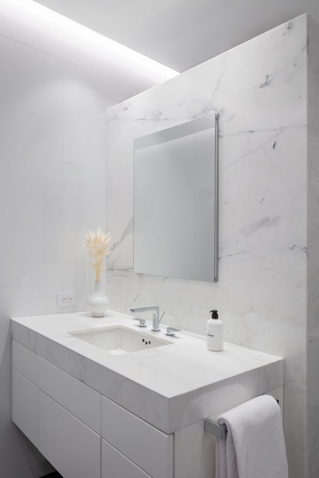 Bright minimalist bathroom vanity with a white marble-look wall and benchtop, integrated rectangular basin, wall-mounted mirror, sleek chrome tapware, and streamlined white cabinetry. A concealed linear ceiling light adds an even, contemporary glow.