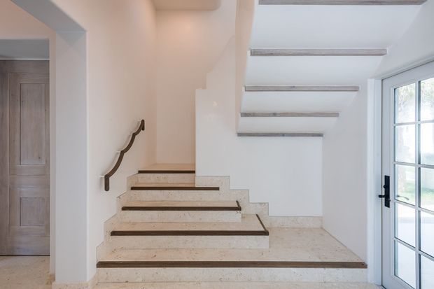 Bright, minimalist stairway with broad terrazzo or stone steps, dark contrasting stair nosings, and exposed stair structure above. White walls and a large black-framed glazed door create an airy contemporary interior. A simple dark handrail runs along the wall, with a timber-panelled door visible in the adjacent opening.