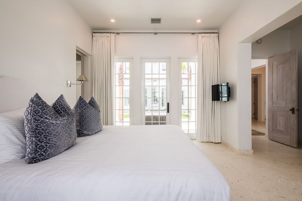 Bright, refined bedroom with a large white bed dressed in crisp white linens and patterned blue-grey accent pillows. Full-height white curtains frame glazed French doors, while a wall-mounted television and a natural timber door are visible to the right. The room has pale walls, recessed ceiling lights, and a light neutral floor, creating an airy, calm atmosphere.