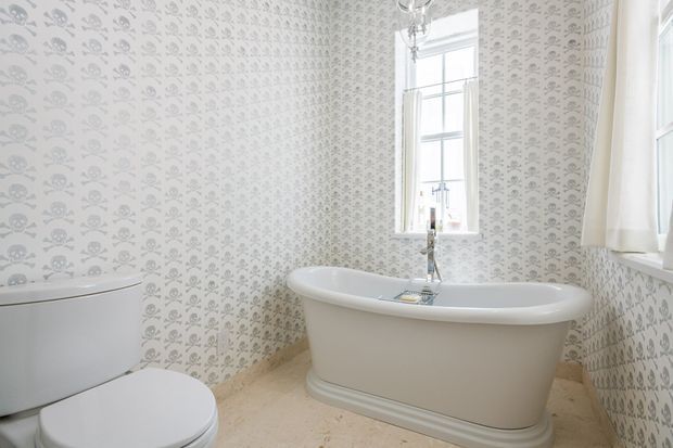Bright traditional bathroom featuring a freestanding white pedestal bathtub, white toilet, chrome floor-mounted bath tap, tall sash windows, and patterned grey-and-white wallpaper with a repeating skull-and-crossbones motif. The floor is a light beige stone or tile, and the room has a refined heritage-inspired character.